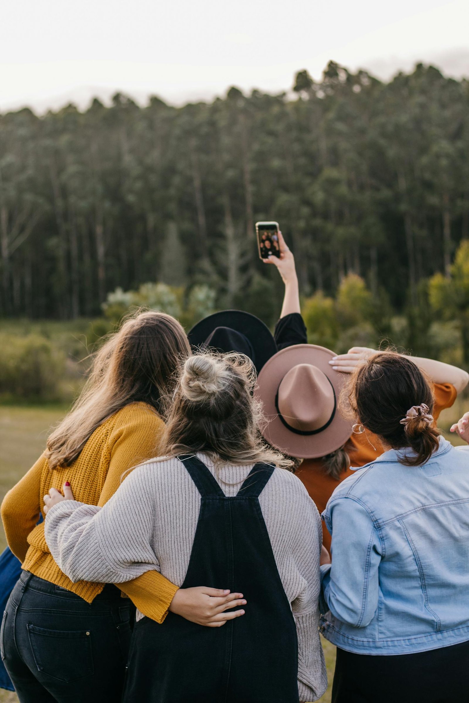 Back view of unrecognizable group of friends taking self portrait on smartphone while standing close to each other near green forest during trip in countryside